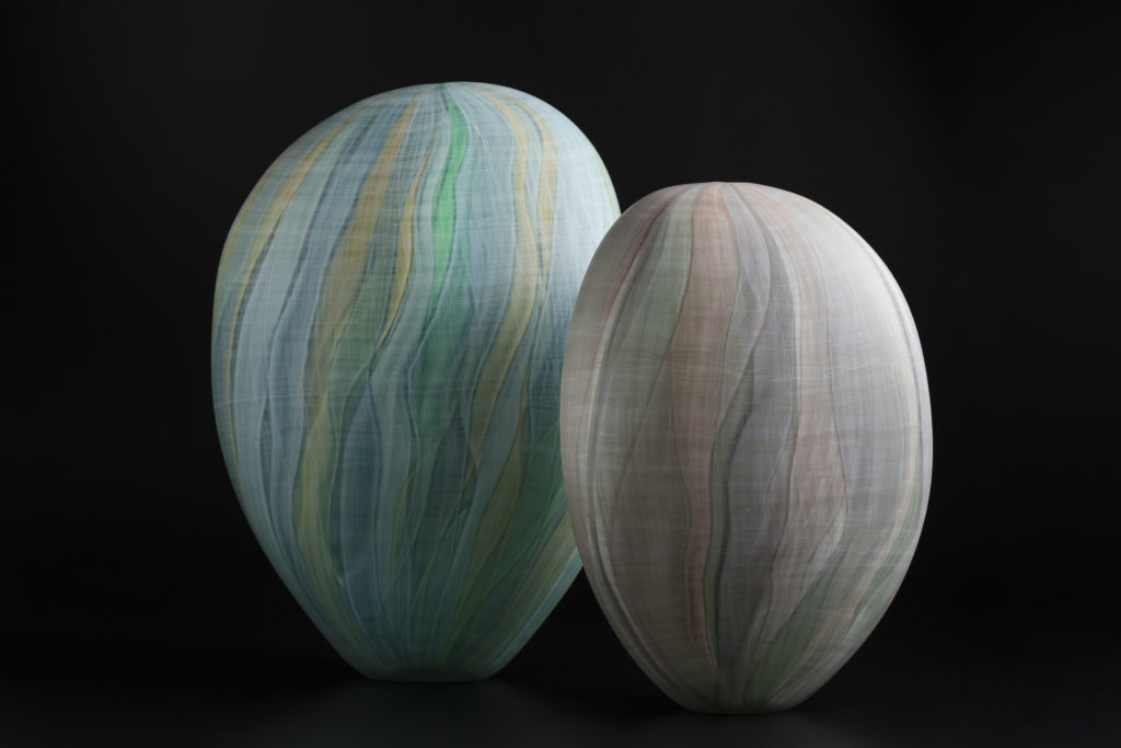 A pair of coloured glass forms standing upright against a black background. The form on the left is slightly larger and is decorated with vertical bands of in green, blue and yellow. The smaller piece ion the right shows bands in dusty pink, grey and green hues.